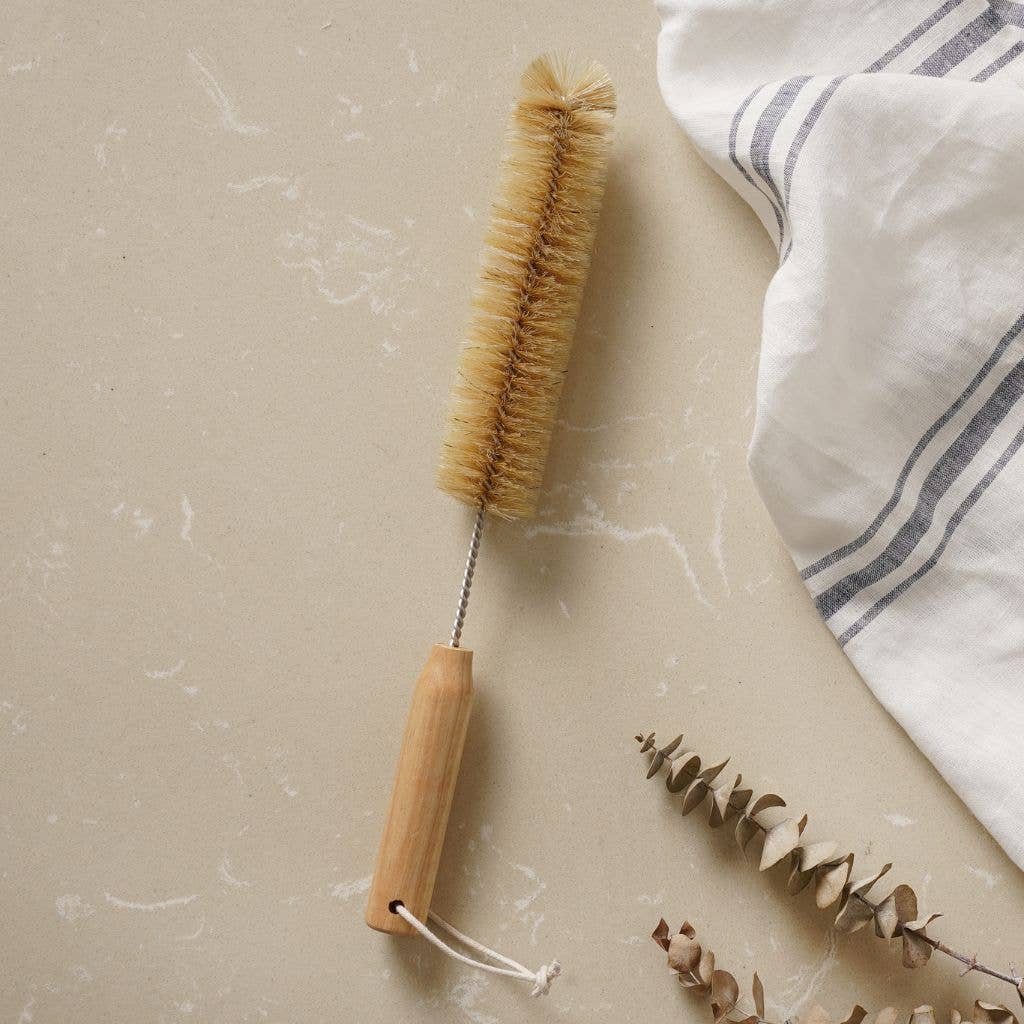 Bottle brush with wooden handle on a marble surface with a white cloth and dried leaves.