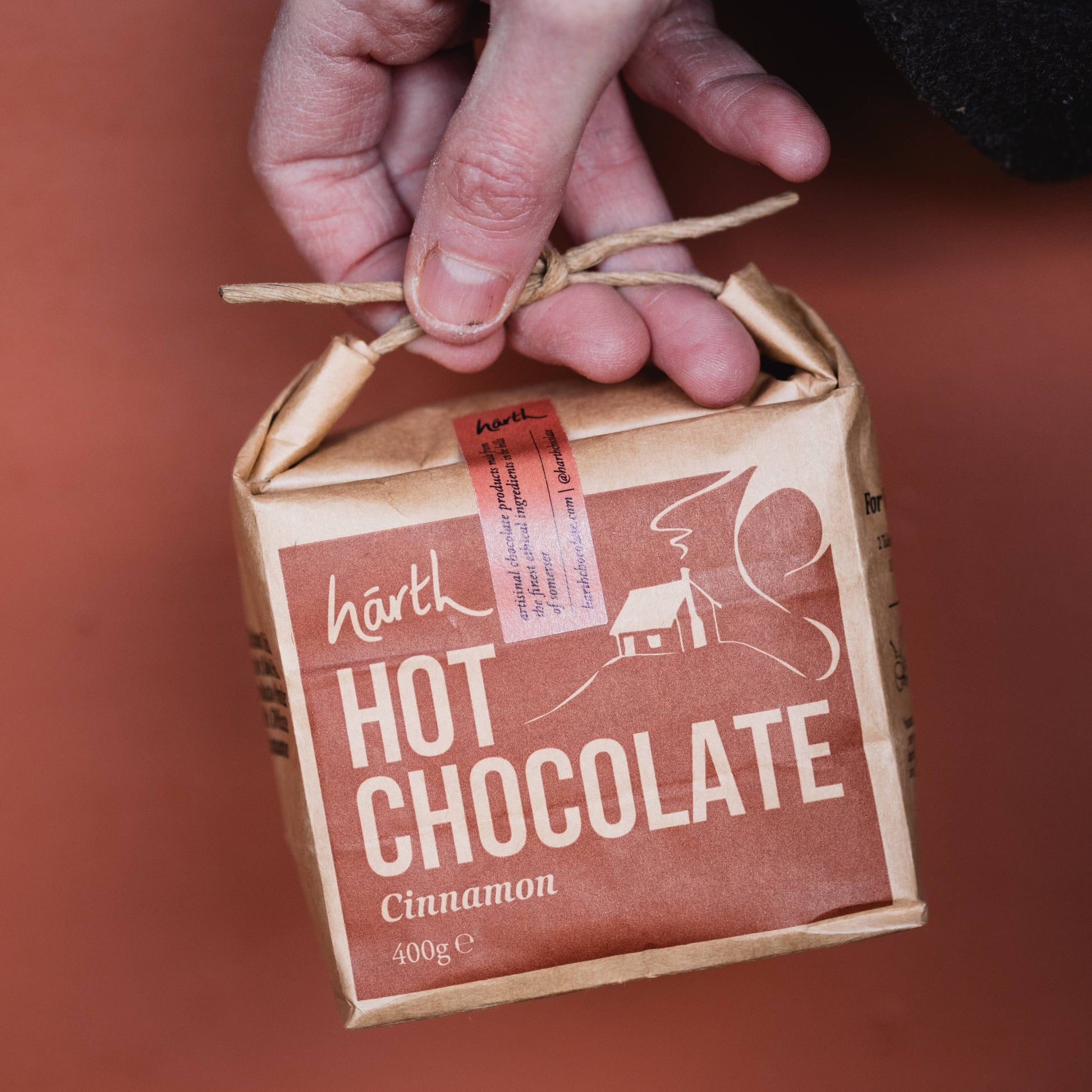 Hand holding a small cardboard box of Hush Hot Chocolate Cinnamon on a red background