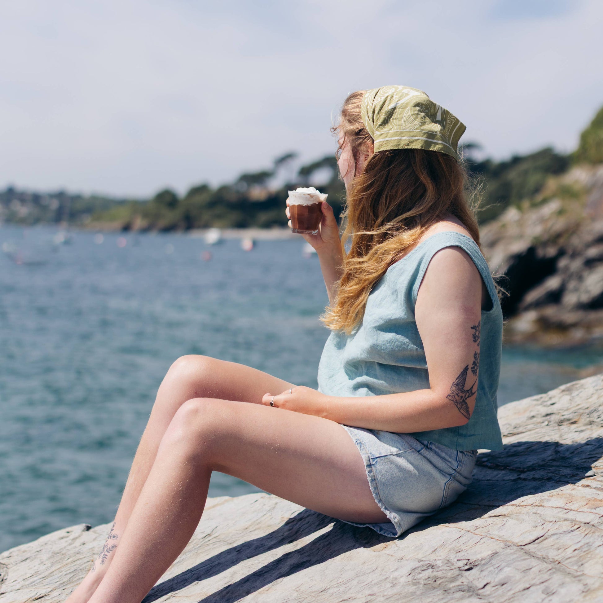 Person sitting by a body of water holding a cup, wearing a light blue sleeveless shirt and denim shorts.