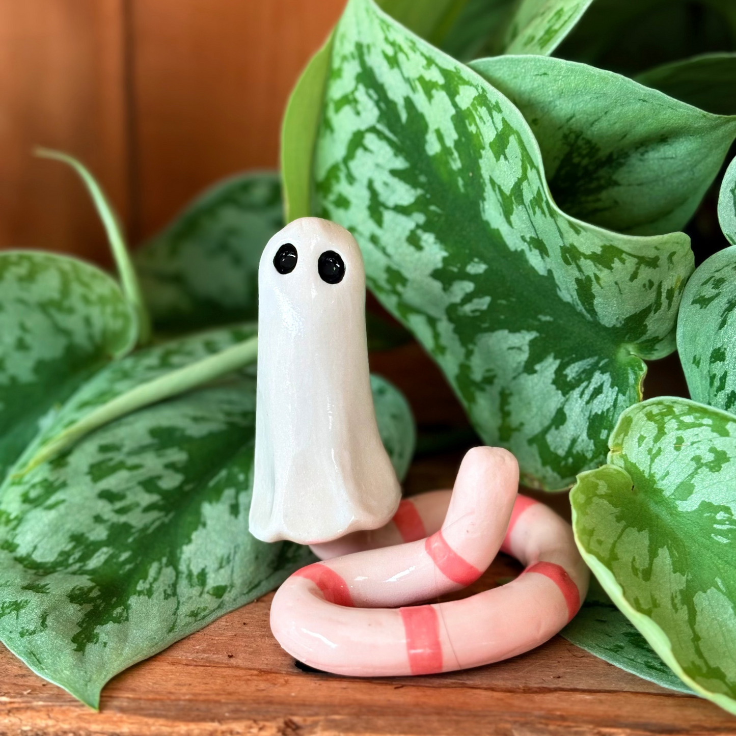 Small figurine of a ghost with a worm-like body among green leaves on a wooden surface