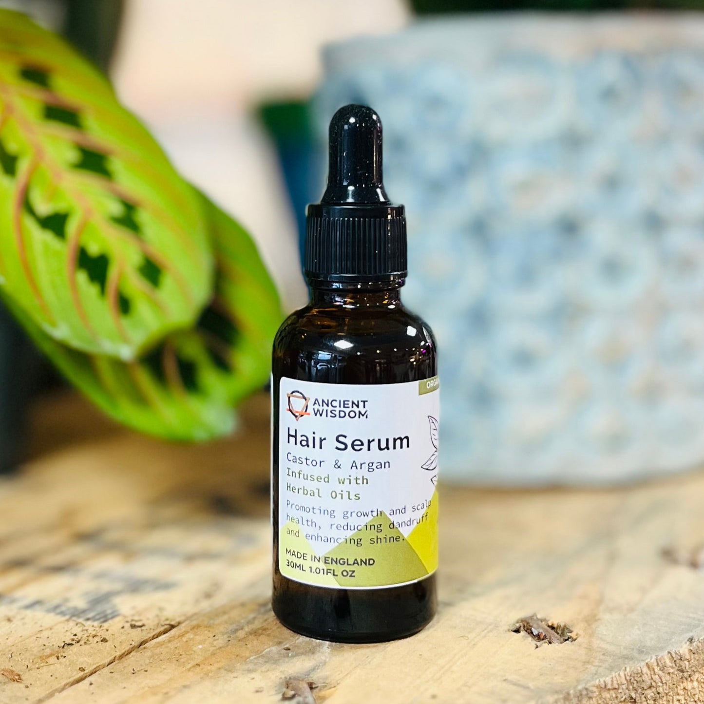 A bottle of Ancient Wisdom Organic Herbal Hair Growth Serum with a dropper cap, placed on a wooden surface with a plant in the background.
