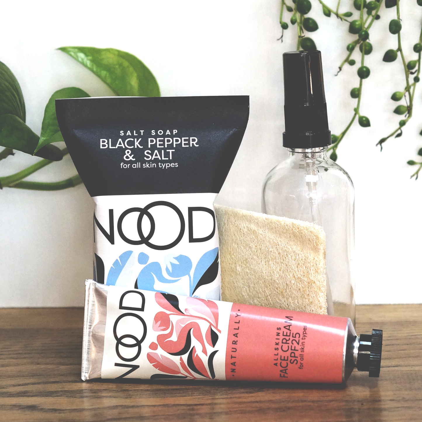 a bar of 'Black Pepper & Salt' Salt Soap, next to a clear glass pump bottle with a black pump top, a loofah and a tube of 'Allskins' Face Cream with SPF 25 lying next to it, all behind a white background with greenery behind it.