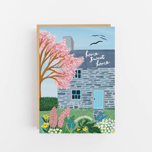 Greeting card with a house, cherry blossom tree, and 'home sweet home' text on a light gray background.