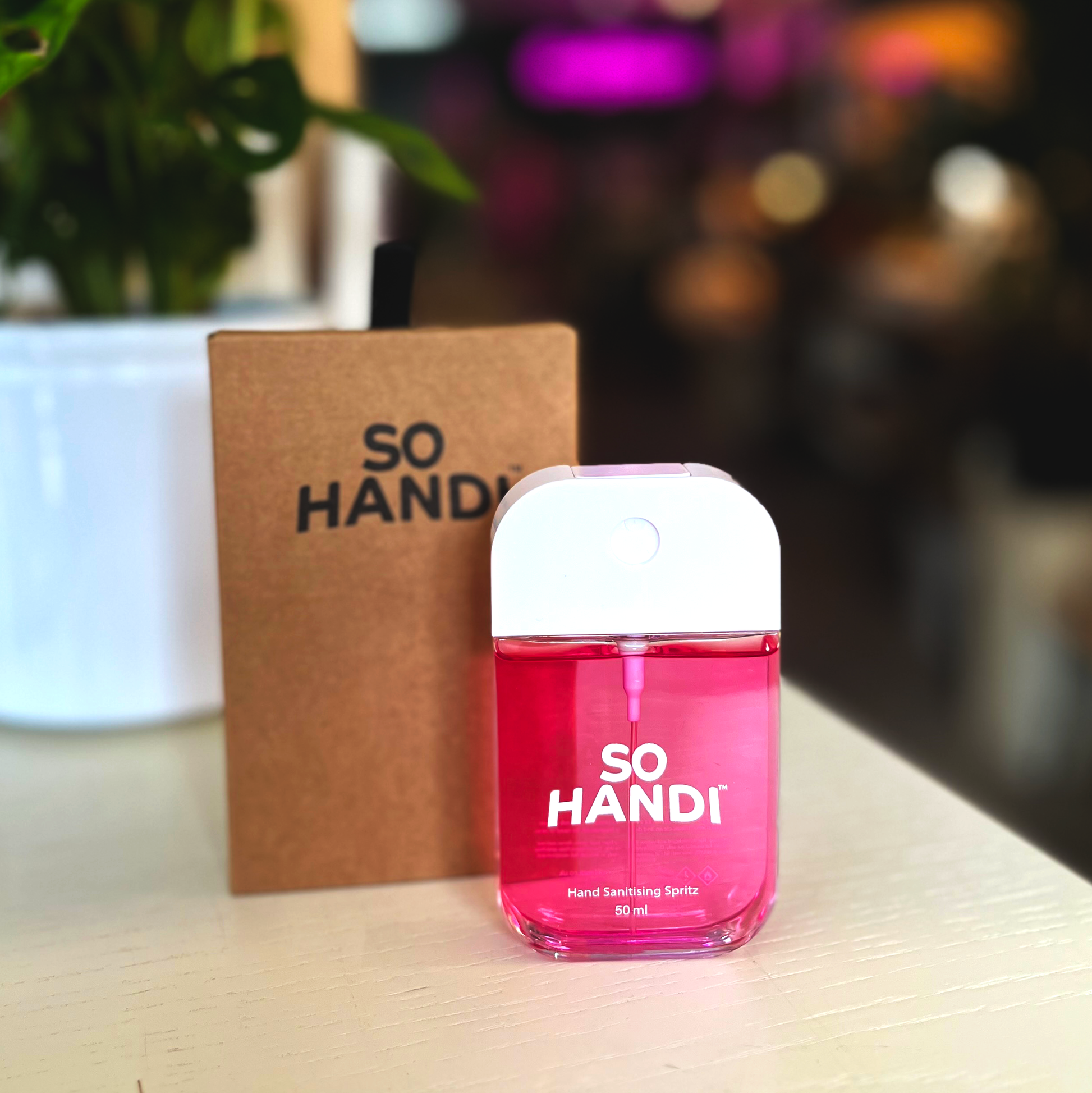 A pink bottle of So Handi hand sanitizer spritzer with its packaging box, placed on a surface with a blurred background.