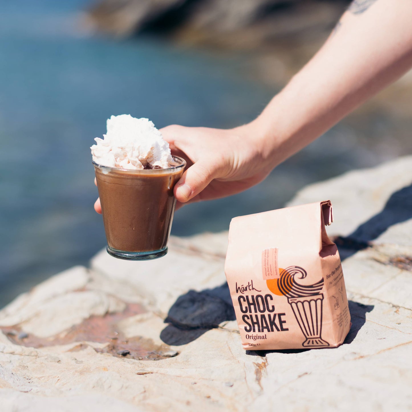 Hand holding a Choc Shake with whipped cream by a scenic coastal background