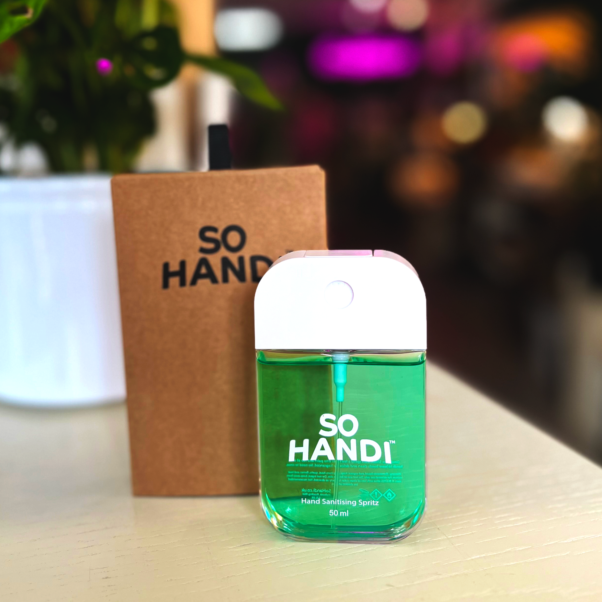 A rectangular spritzer bottle of green hand sanitiser, stood in front of its recyclable packaging. The product is branded as 'So Handi'.