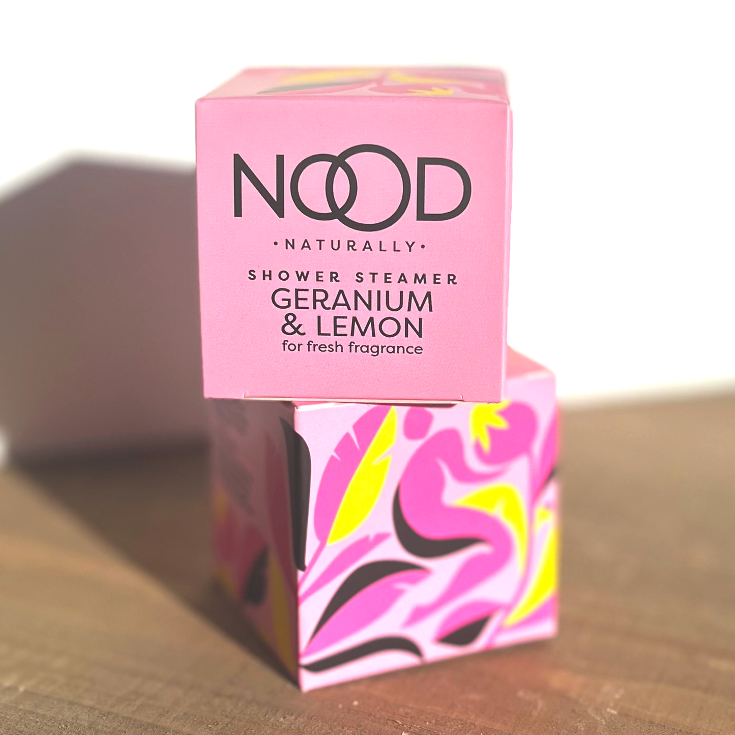 Two small pink boxes stacked, with the branding of 'NOOD' on them alongside an abstract floral pattern. They have 'Shower Steamer Geranium & Lemon' written on them.
