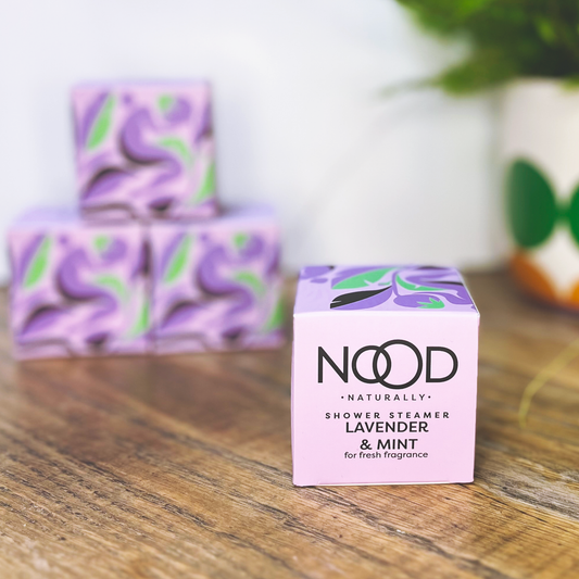 A small square lilac box, containing a 'Lavender & Mint Shower Steamer' with 'NOOD Naturally' written on it. It also has an abstract, almost floral pattern on it.