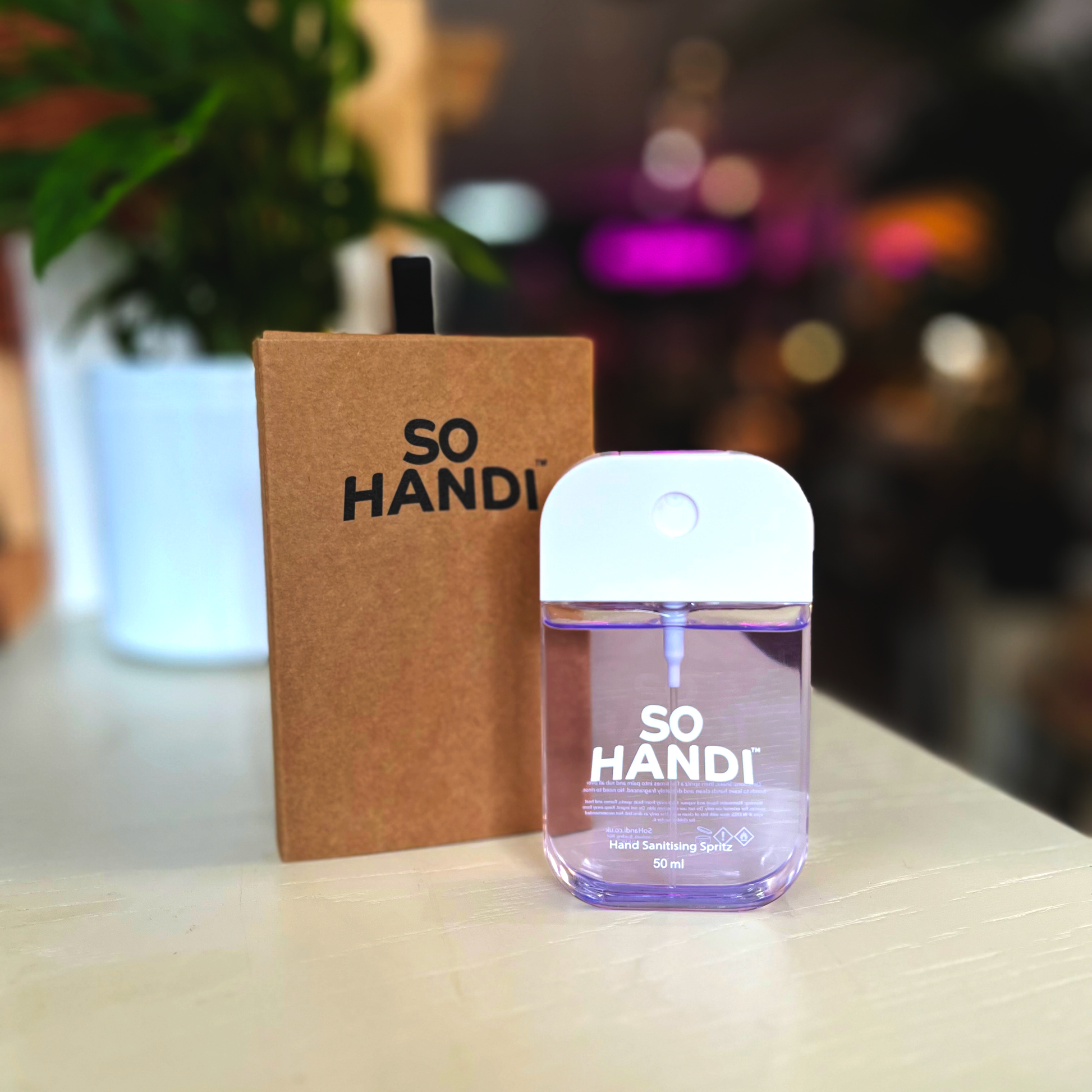 A rectangular hand sanitiser spritzer, filled with purple sanitiser. It stands in front of a box branded So Handi.