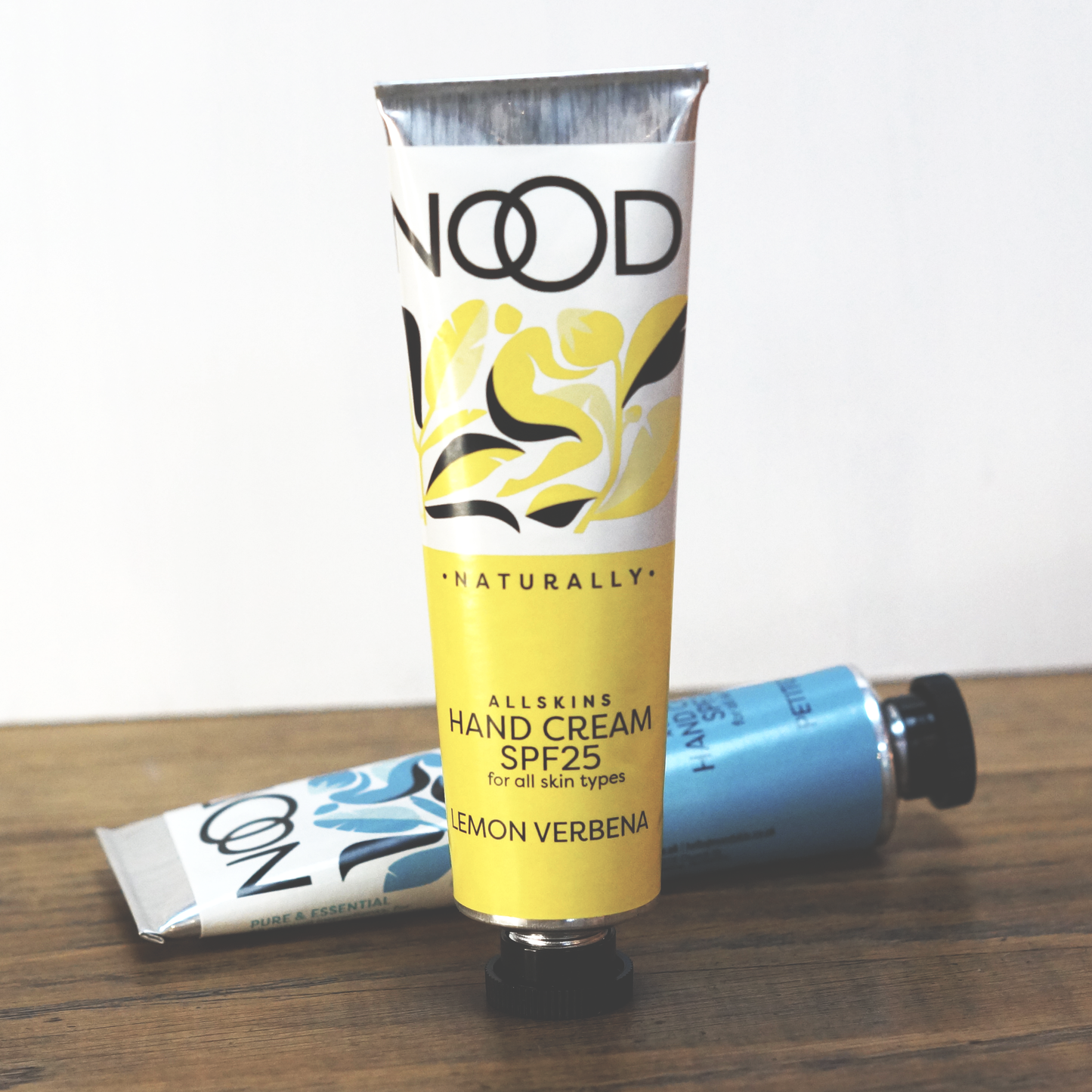 A tube of NOOD Stores Lemon & Verbena Hand Cream with SPF 25, placed on a wooden surface, with its cap removed and a second tube partially visible in the background.