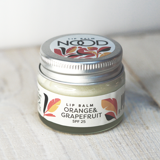 A glass jar of NOOD brand lip balm with orange and grapefruit flavor, featuring a colorful label with floral designs.