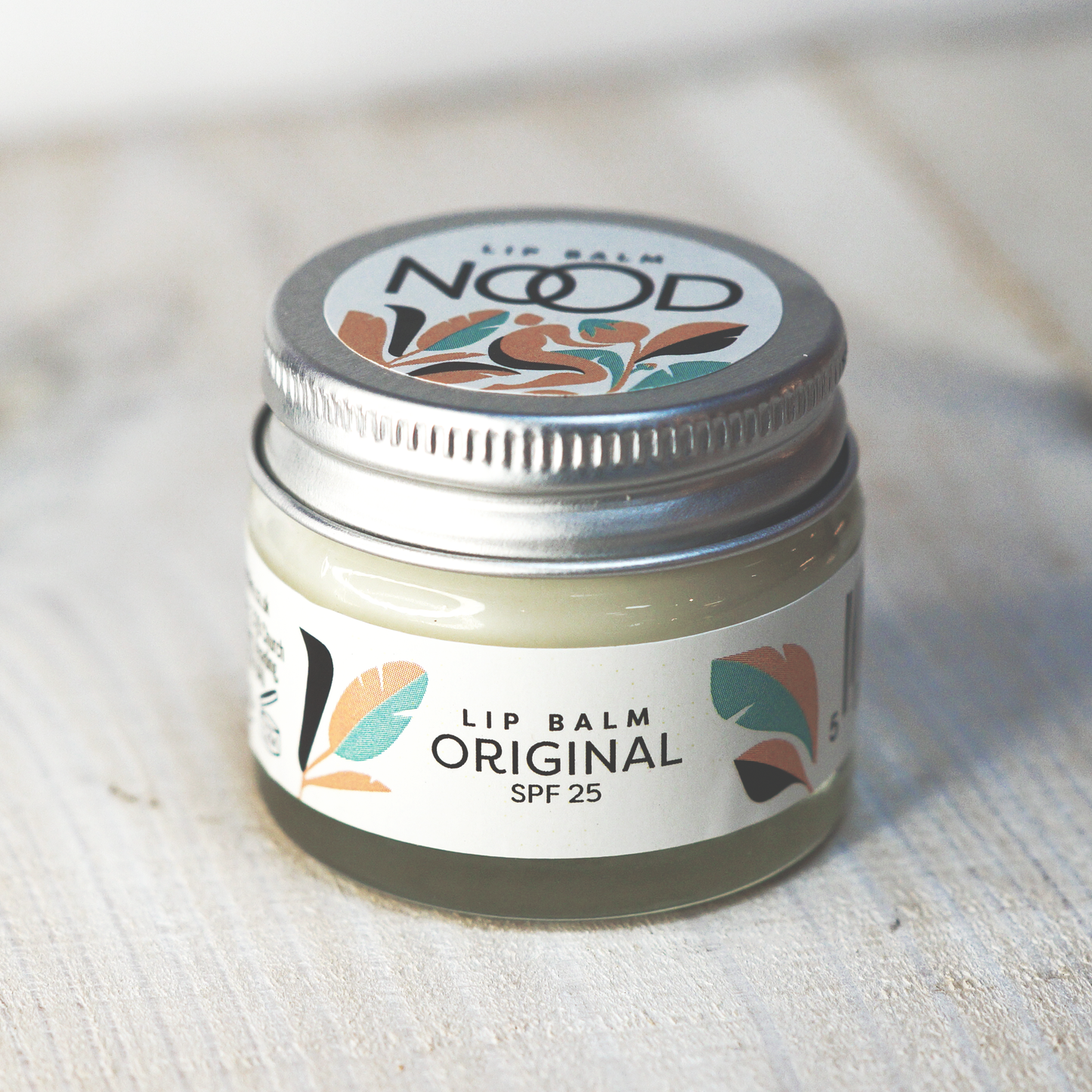 A glass jar of NOOD Original Lip Balm SPF 25, placed on a wooden surface. The label shows the brand name and product details.