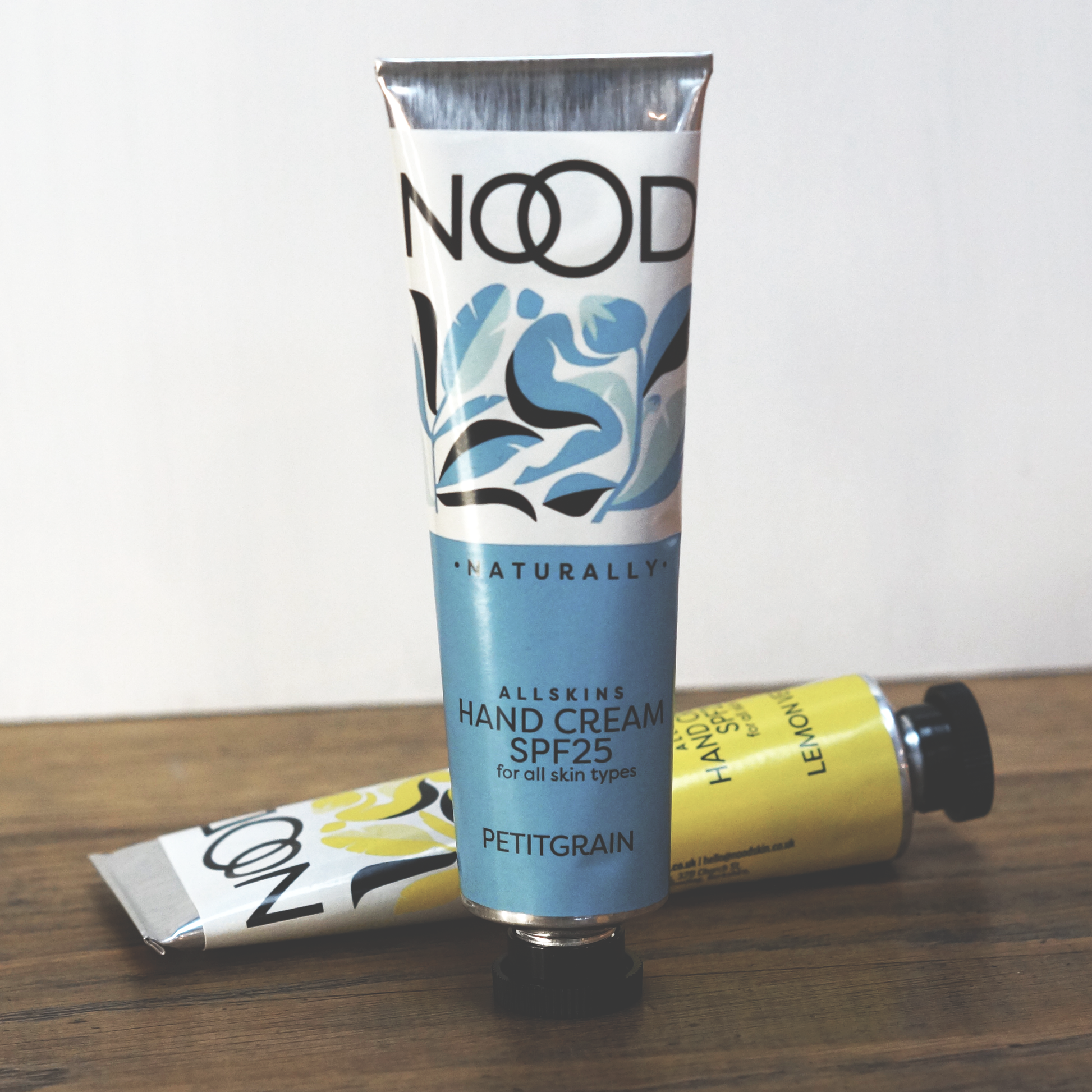 A tube of NOOD naturally Petitgrain Hand Cream with SPF 25, placed on a wooden surface, with its packaging predominantly white and blue with floral and leaf patterns.