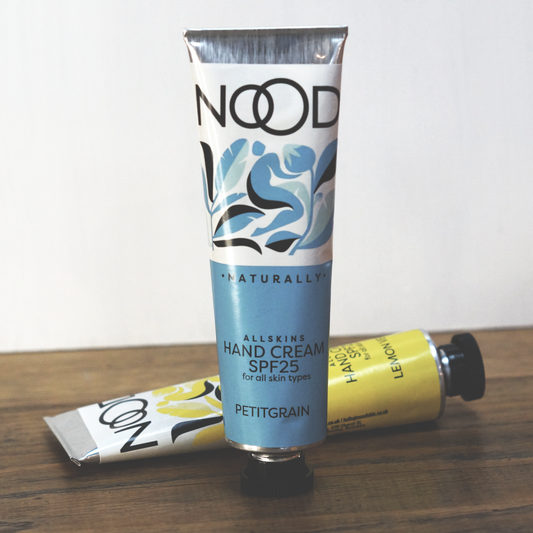 A tube of NOOD naturally Petitgrain Hand Cream with SPF 25, placed on a wooden surface, with its packaging predominantly white and blue with floral and leaf patterns.