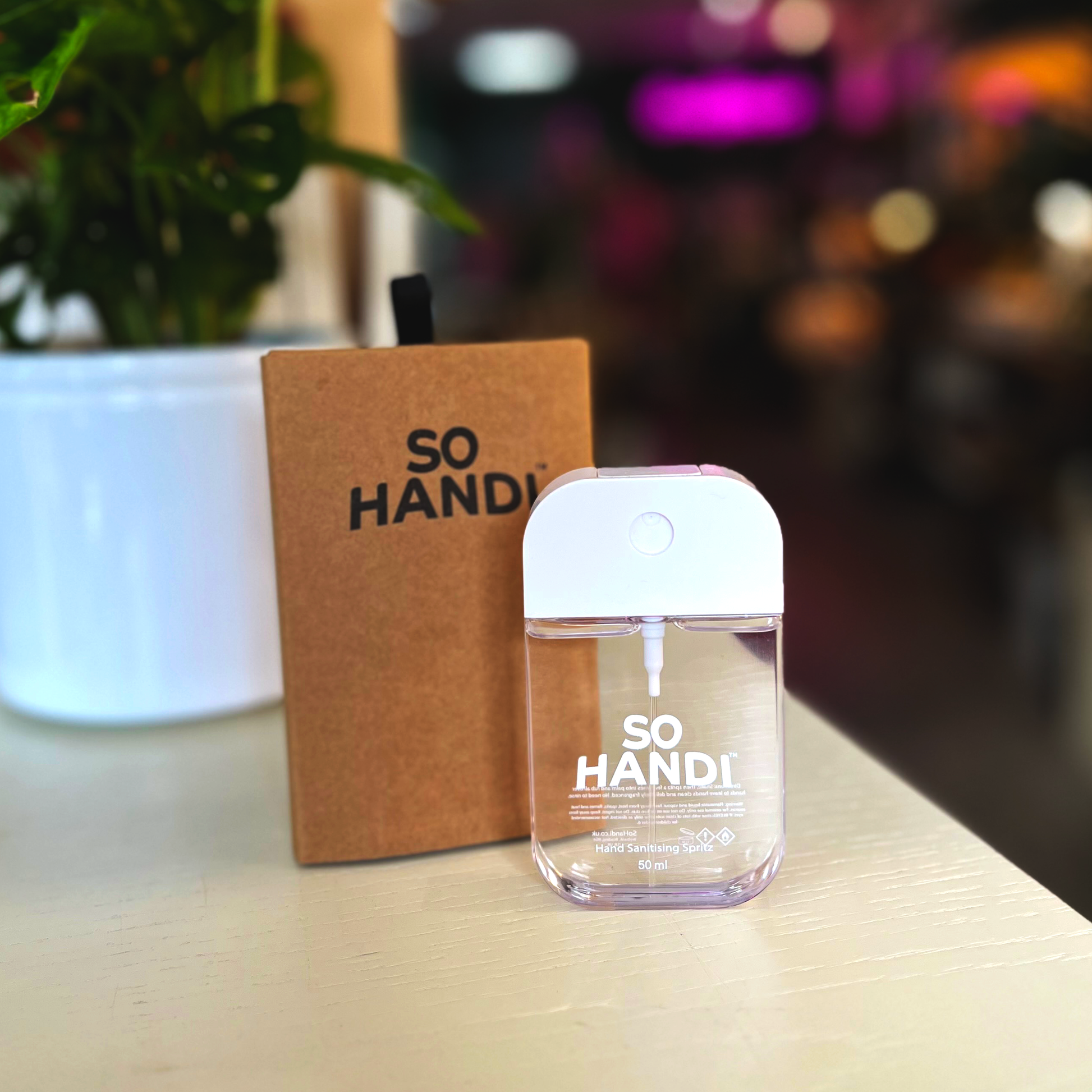 A rectangular sanitiser spritzer stands in front of a brown box that says So Handi.