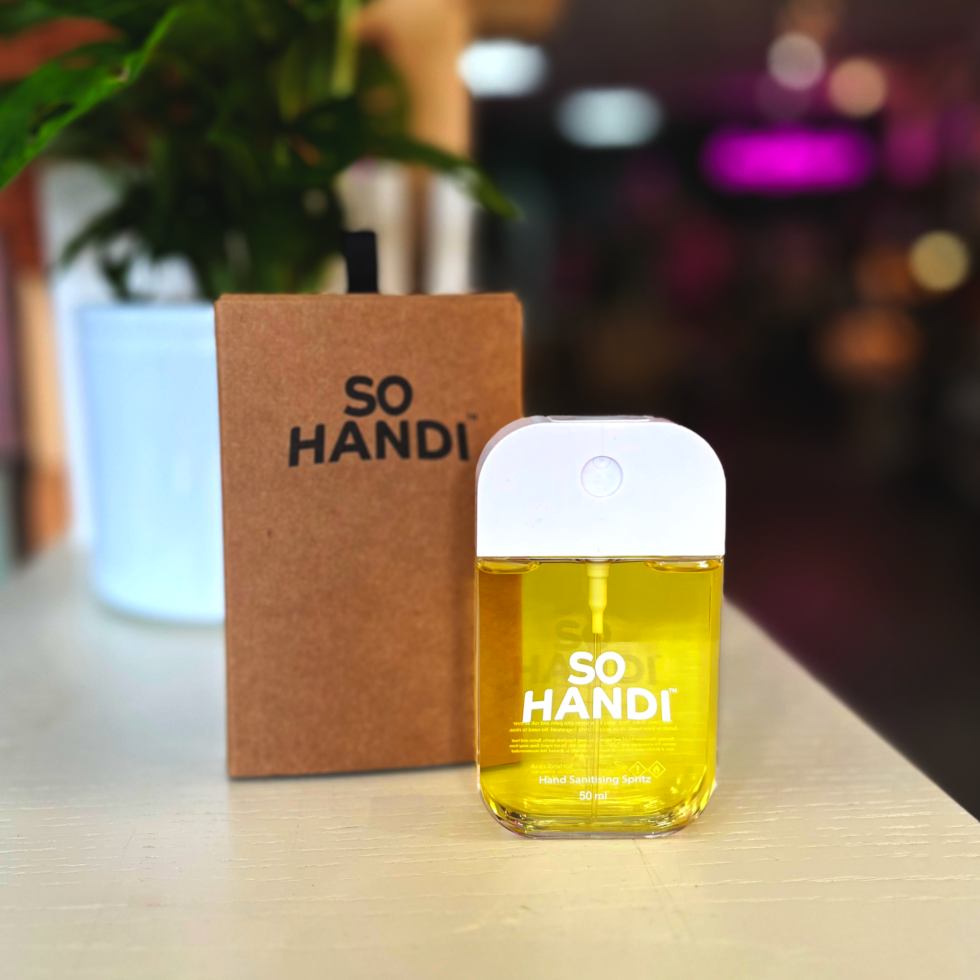 A rectangular hand sanitiser spritzer stands in front of its brown box marked 'So Handi'. This spritzer is filled with yellow hand sanitiser.