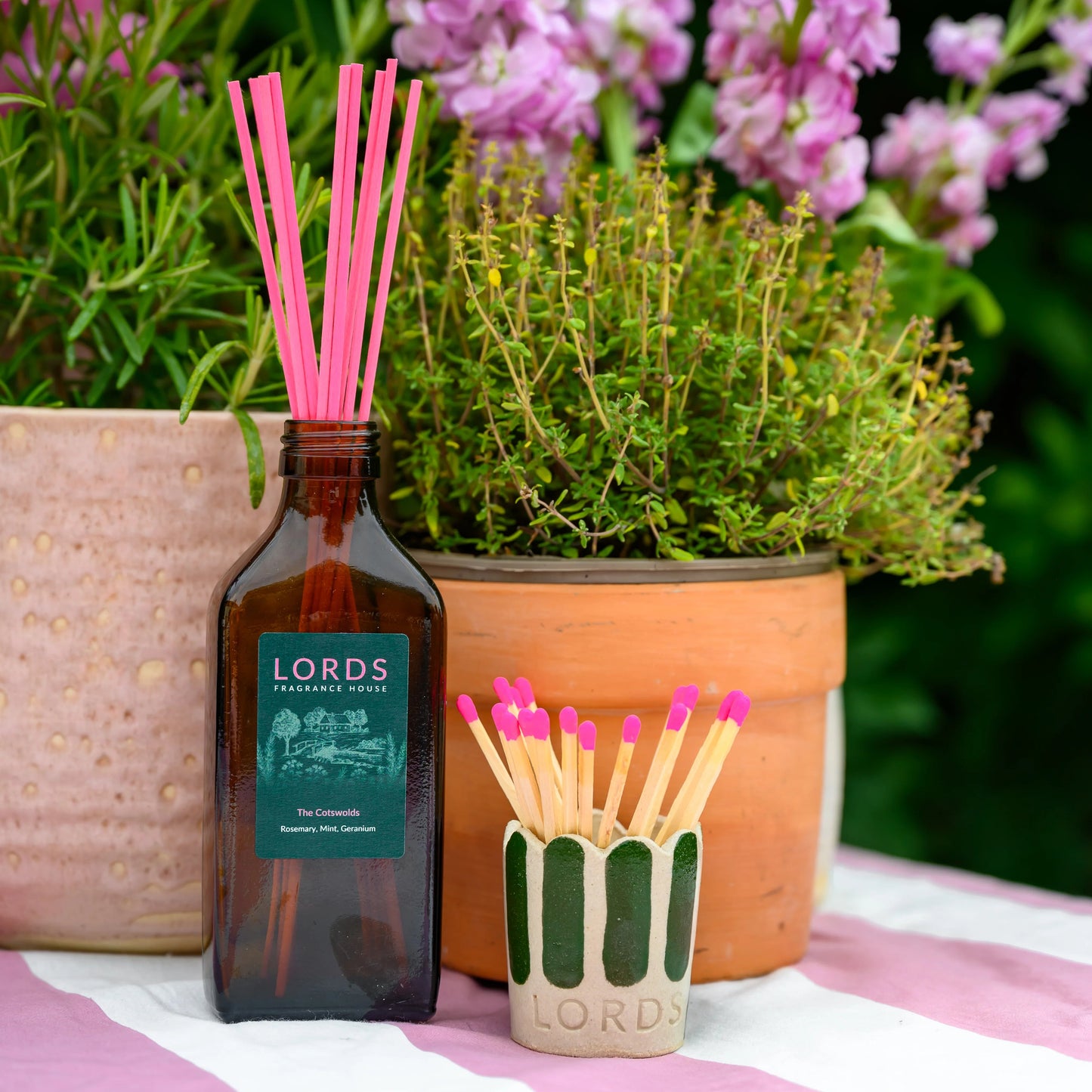 Bottle of Lords with pink reeds and matches on a striped surface with flowers in the background