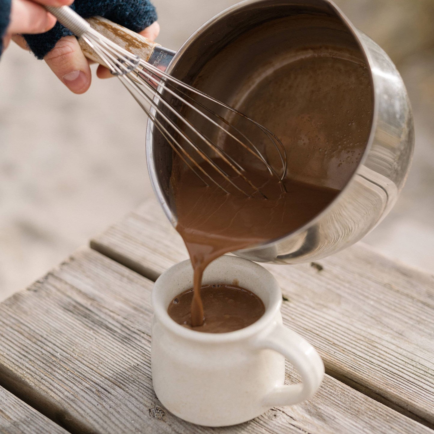Person pouring chocolate mixture from a metal pot into a white mug on a wooden surface.