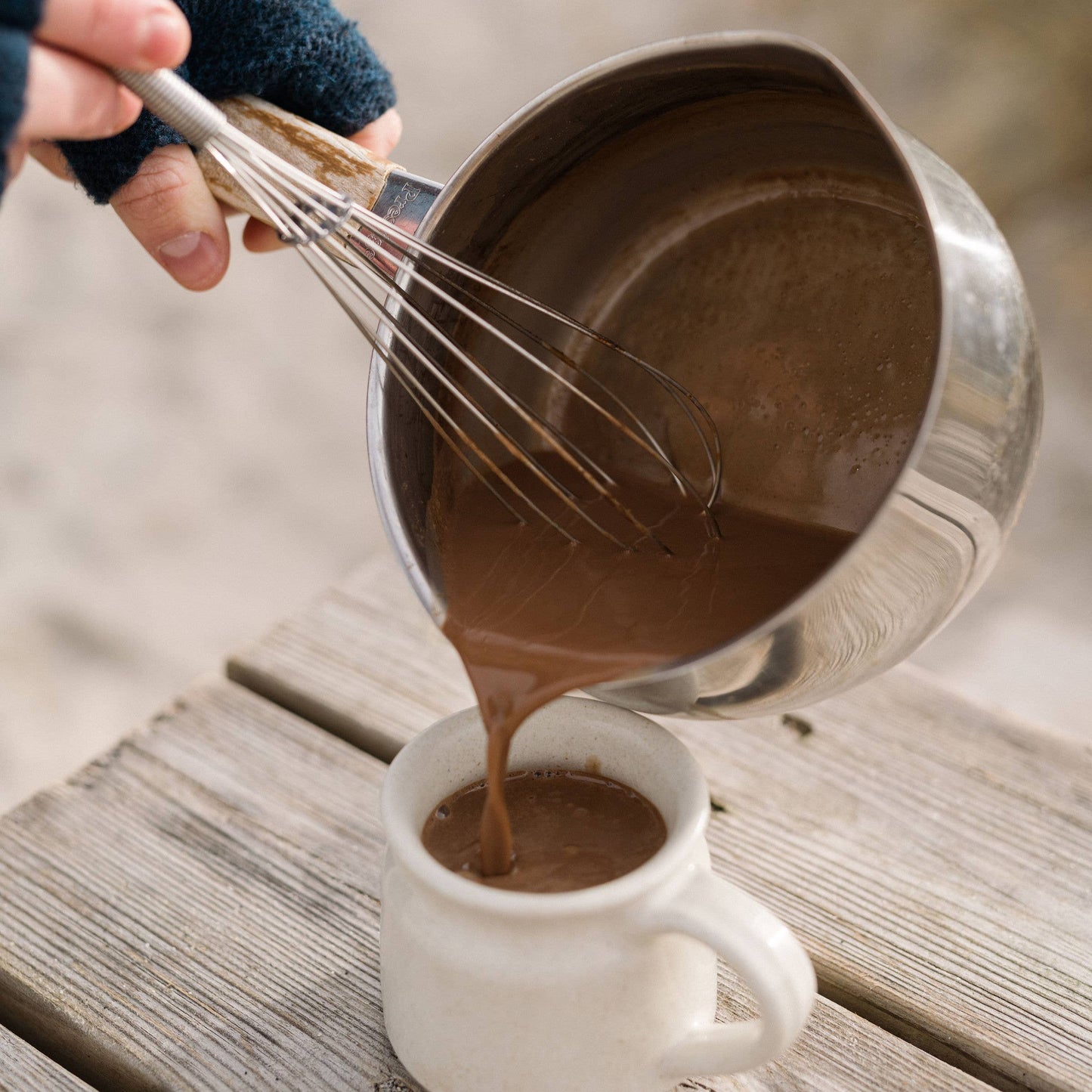 Person pouring chocolate mixture from a metal pot into a white mug on a wooden surface.