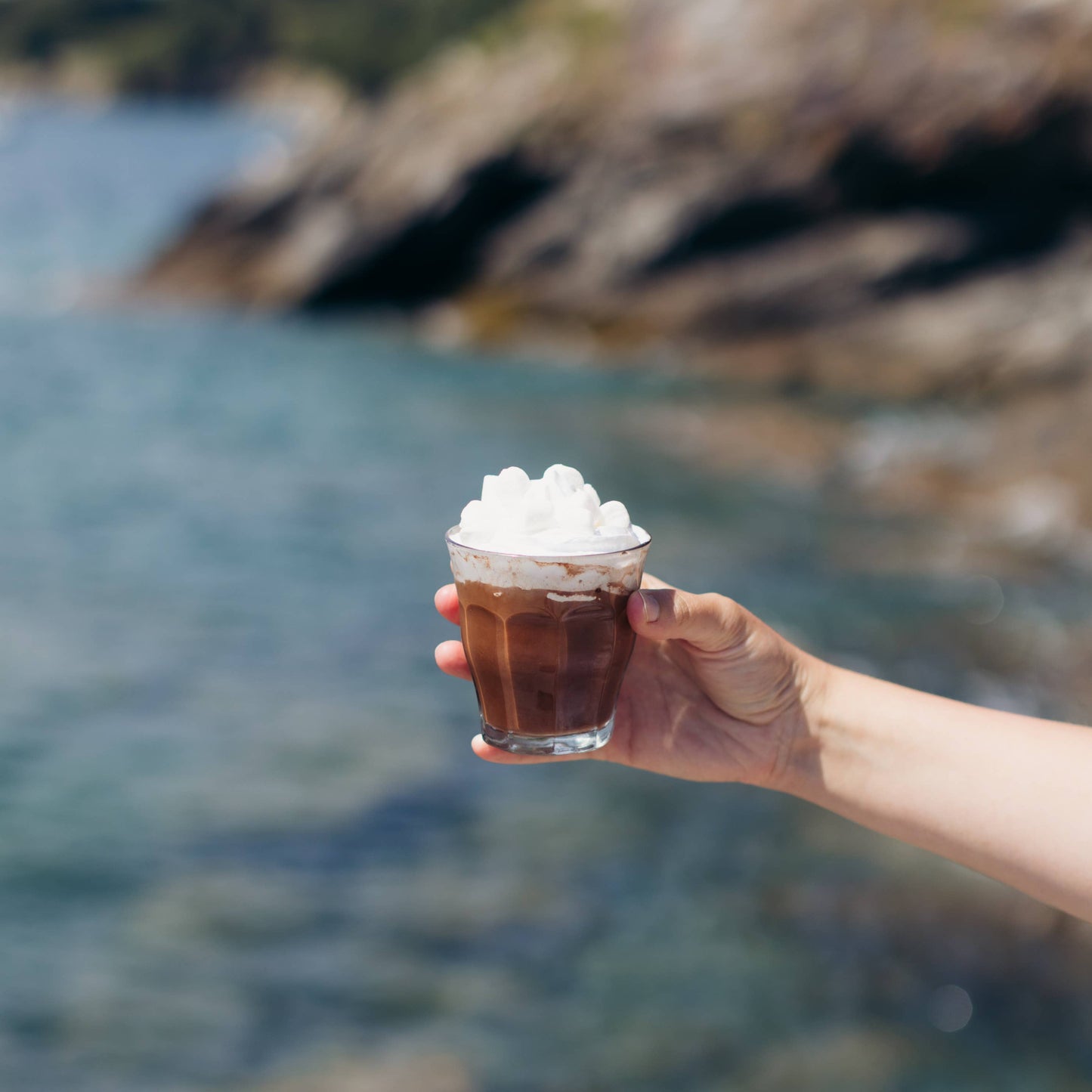 Hand holding a glass of iced coffee with whipped cream against a blurred natural background
