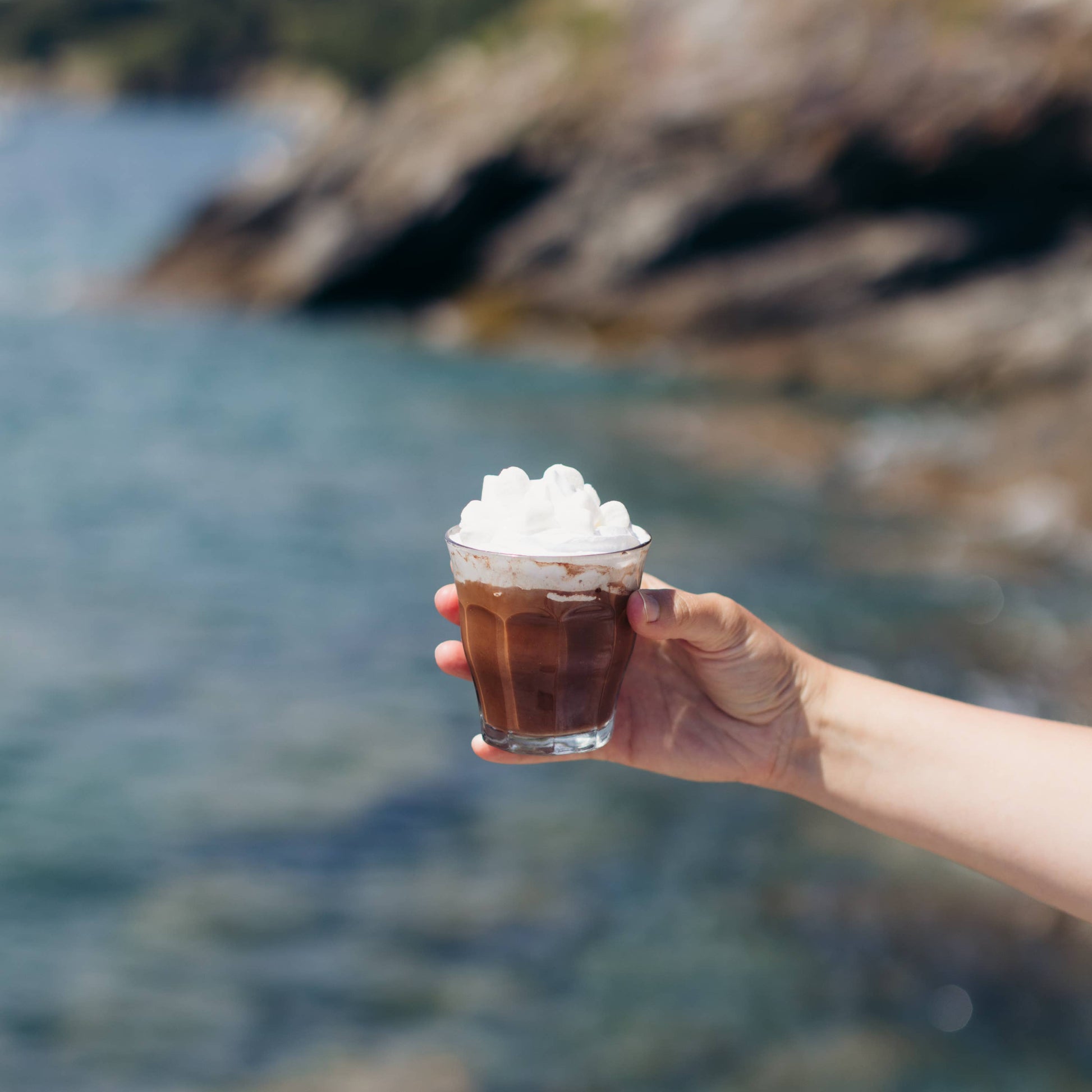 Hand holding a glass of iced coffee with whipped cream against a blurred natural background