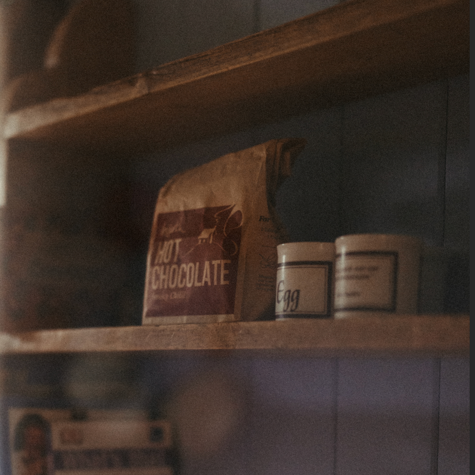 Shelves with a bag of hot chocolate, a jar labeled 'Egg', and a box of 'Flowers' on a tiled wall.