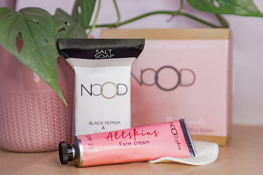 A slightly blurred background with a nice plant hanging over. The black and white packaged soap sits in front of the outer packaging box, wrapped in a light pink band, and the Allskins Moisturiser tube, in a pink label, lays in front of the soap resting on a single bamboo makeup pad 