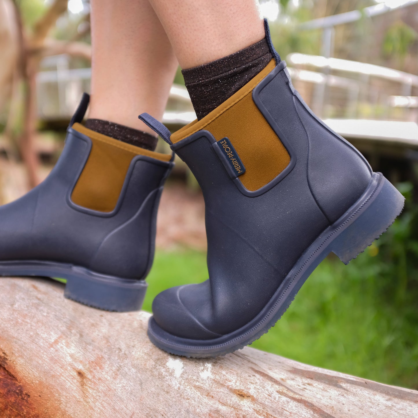 Navy rubber boots with brown accents worn by a person on a log outdoors