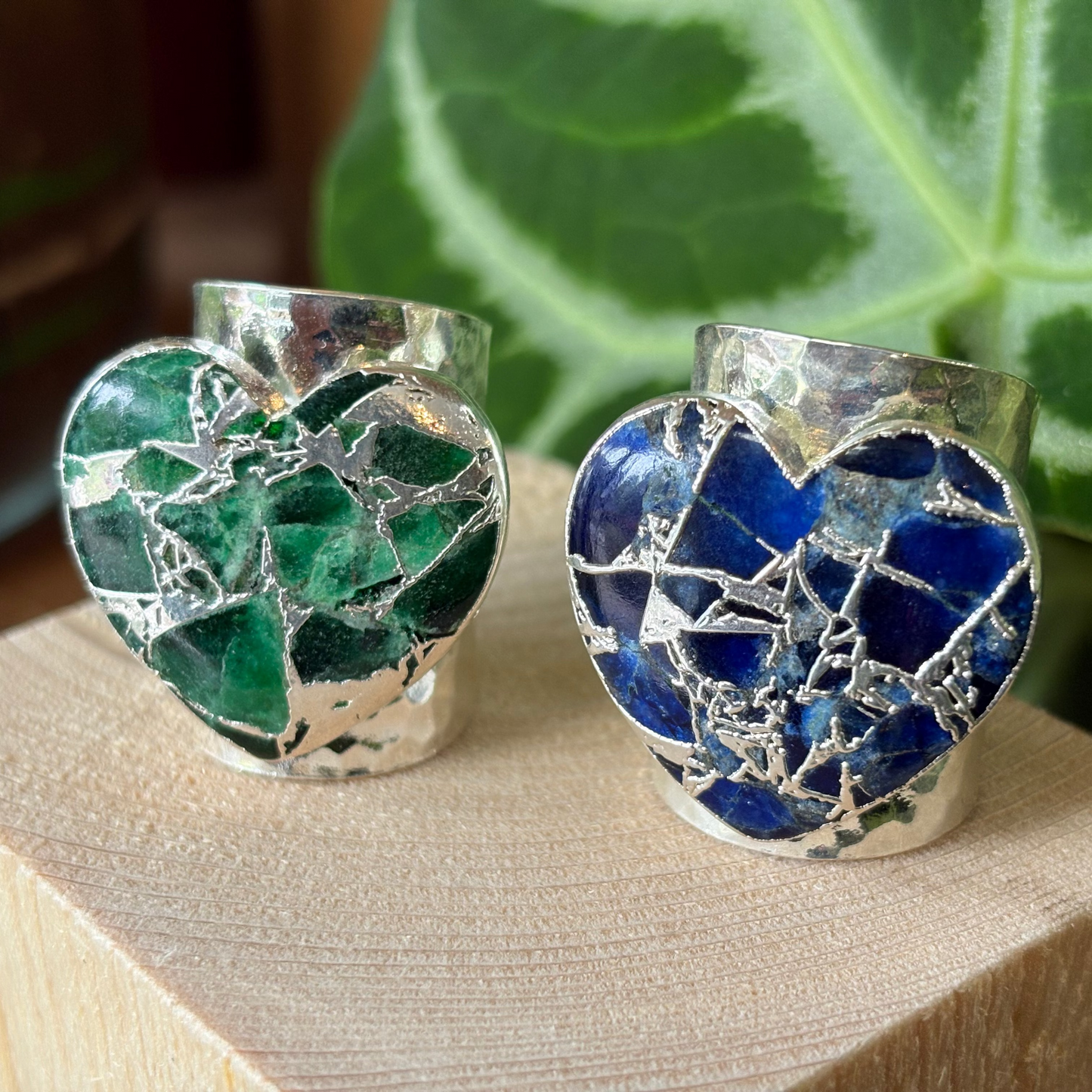 Two heart-shaped rings with green and blue mosaic designs on a wooden block with a plant in the background.