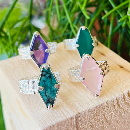 Four colorful gemstone rings on a wooden surface with a green leafy background