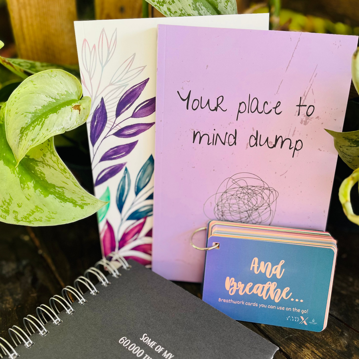 Notebooks and a small book with motivational text on a wooden surface with plants in the background