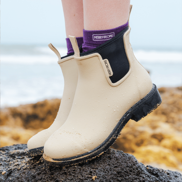 Beige rubber boots with black soles on a rocky surface, wearing purple socks.