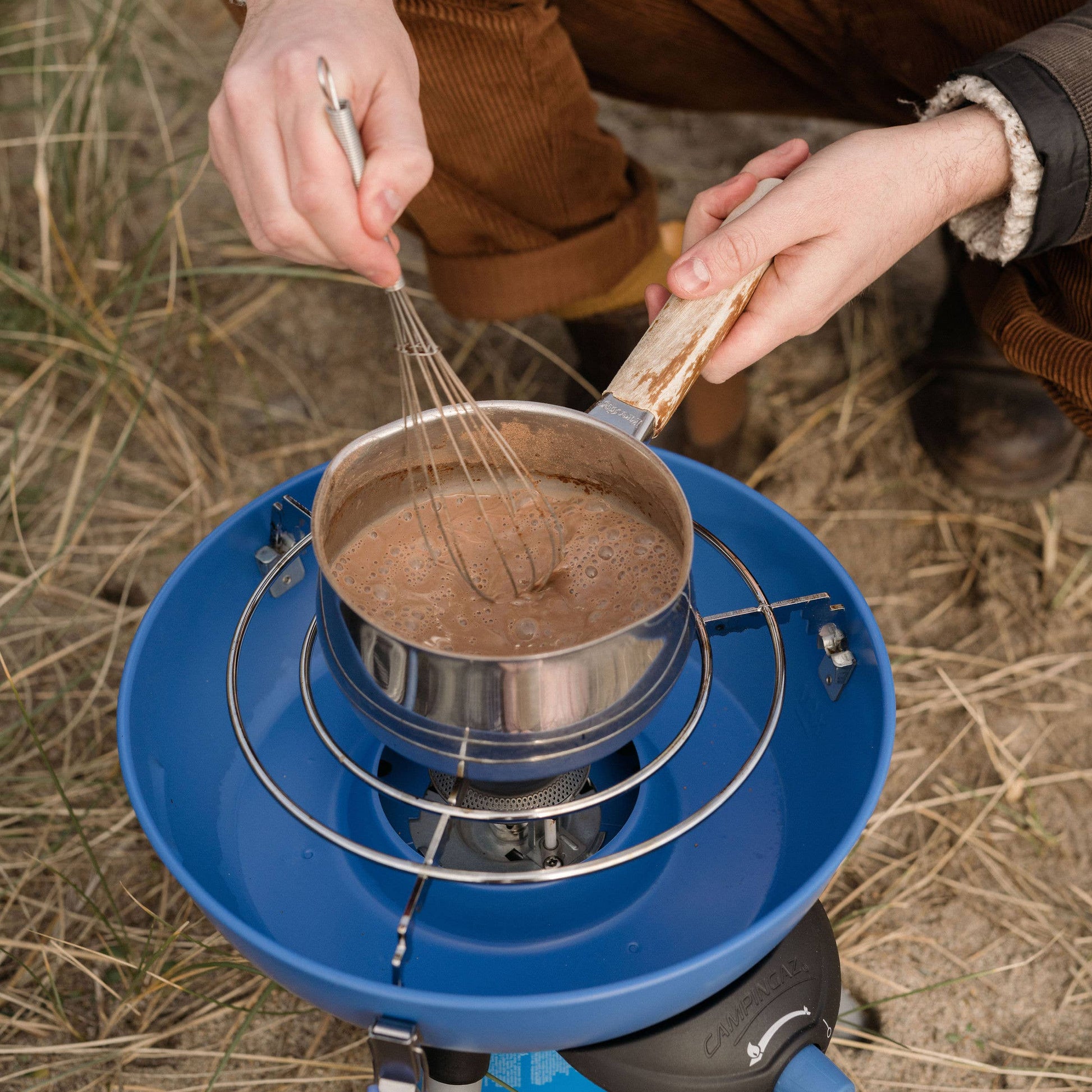 Person cooking outdoors using a portable stove with a pot and whisk
