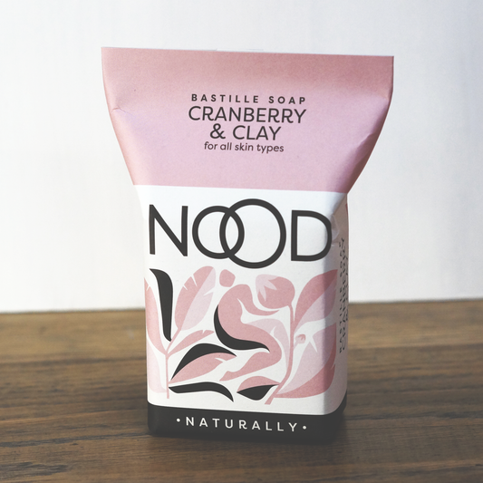 Packaged bar soap with 'NOOD' branding, 'CRANBERRY & CLAY' label, and 'NATURALLY' text, indicating it's for all skin types.