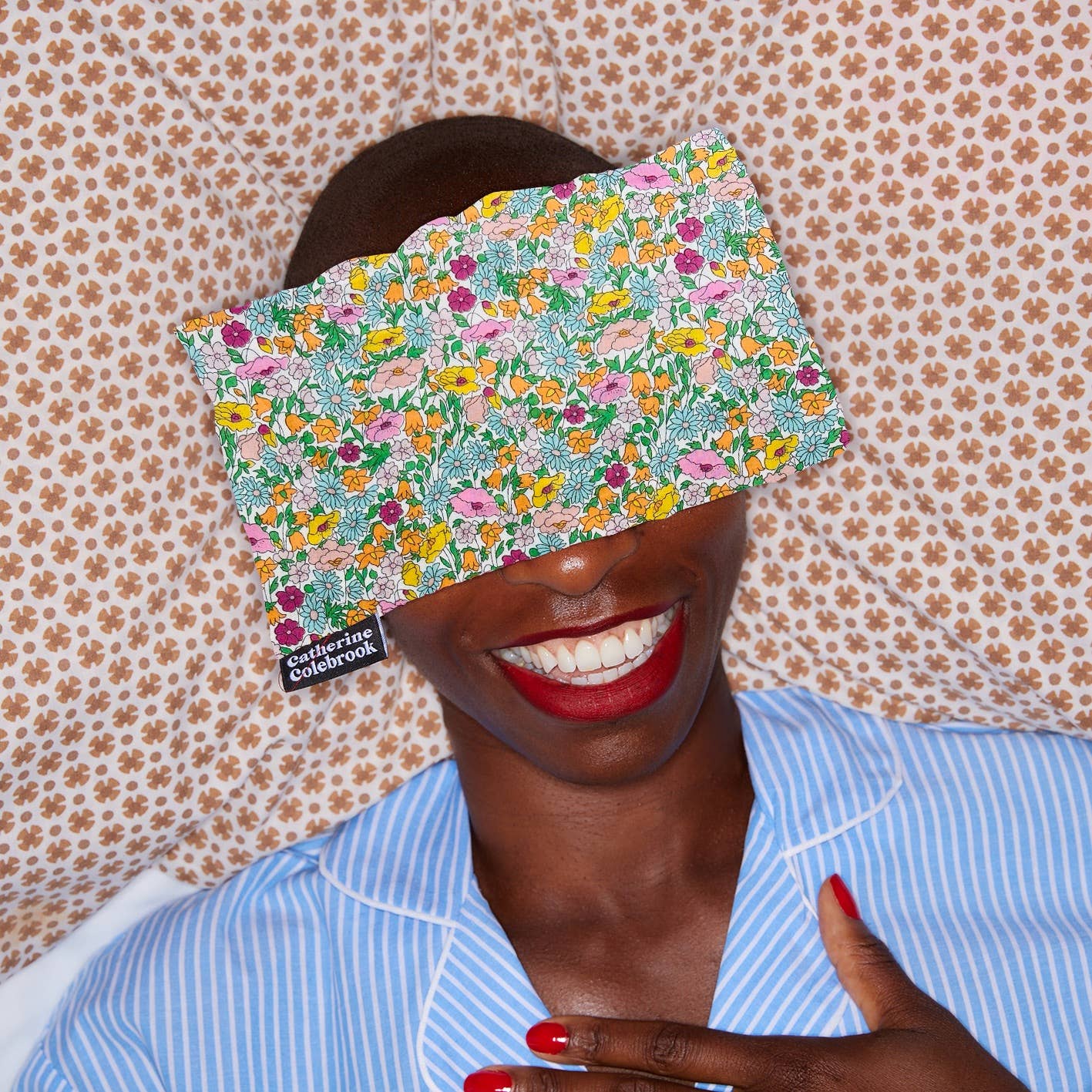 Person wearing a floral sleep mask with a pattern of flowers and text, against a floral wallpaper background.