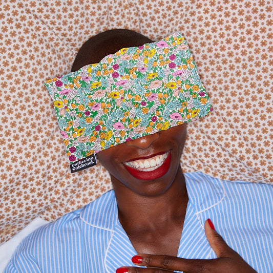 Person wearing a floral sleep mask with a pattern of flowers and text, against a floral wallpaper background.