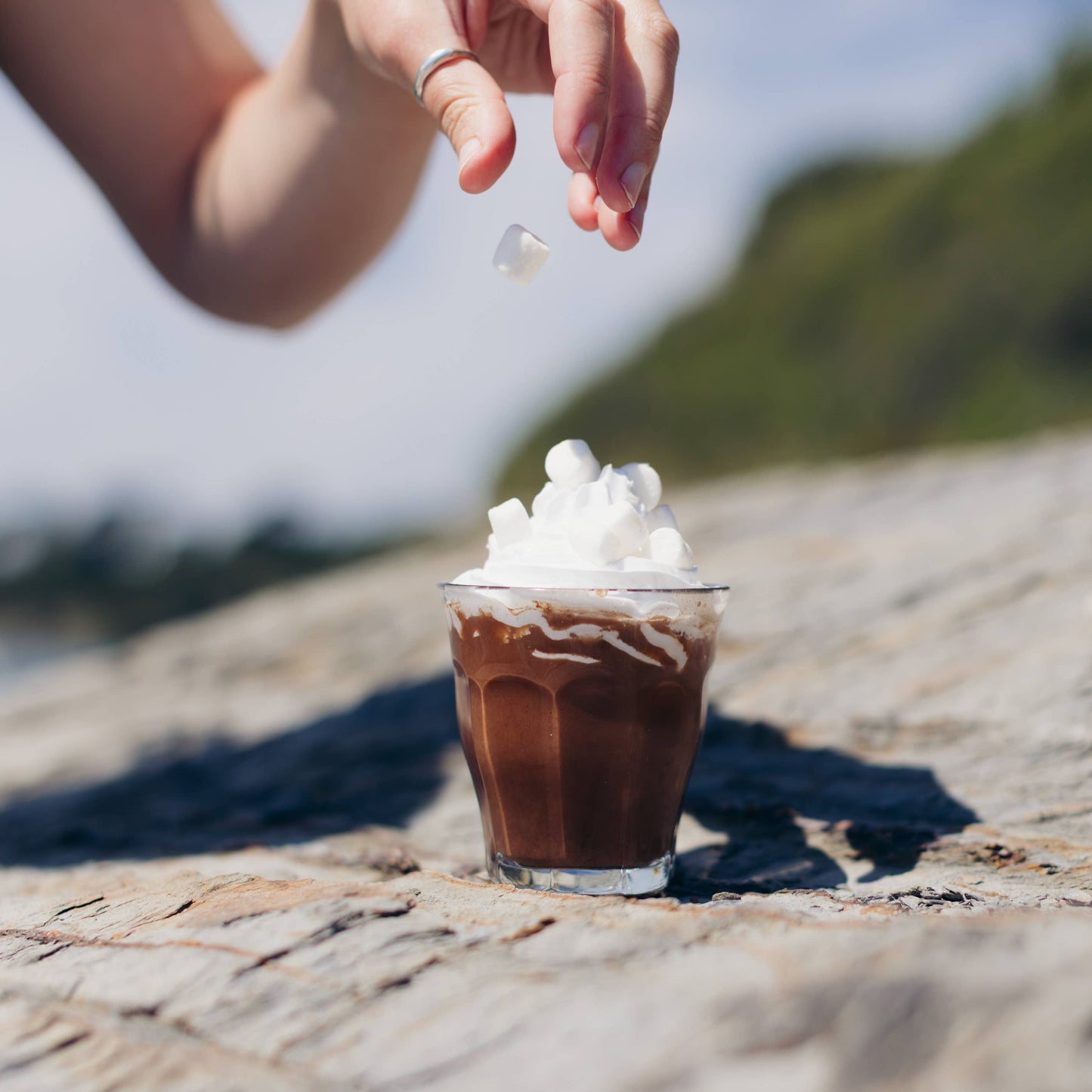 Hand sprinkling a white substance onto a small glass of iced coffee with whipped cream on a stone surface.